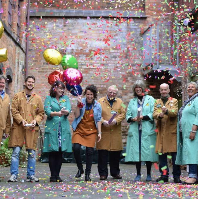 A line of 8 people wearing brown or green workcoats standing in the courtyard of the Coffin Works in Birmingham. Confetti is in the air and 2 people are holding balloons. Everyone is smiling. Tonia is 3rd person from the left wearing a green coat, with mid-length brown hair. 