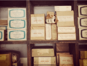 2 wooden shelves with small brown boxes labelled as Coffin Screws and Nickel Plated Handles. Image taken at the Coffin Works in Birmingham. 