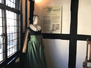 Image shows a mannequin wearing a Tudor style green gown, black kirtle, white ruff and black coif, stood in front of a display board entitled "Who is Margery?". 